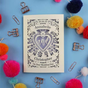A personalised laser cut wedding card with floral filigree papercut design. Card has a space for the date at the top, a banner with a name, a heart shape with the word 'and' and another banner for the second person's name. Card says on your wedding day at the bottom.