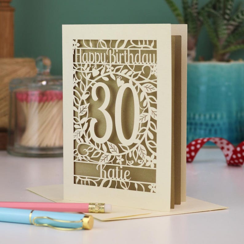 A laser cut 30th birthday card, with "Happy Birthday" text at the top, the number 30 in the centre with a floral and leaf cut out border, and personalised with a name underneath. Card is cream with a gold leaf paper insert.