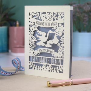 A gorgeous new baby card depicting a stork carrying a bundle surrounded with leaves, flowers and clouds. A banner saying Welcome To The World across the top,  and a second banner below with the baby's date of birth and their name under that. Shown standing on a white desk with stationery items in the fore and background. Its cut from cream cardstock and has a silver insert paper.