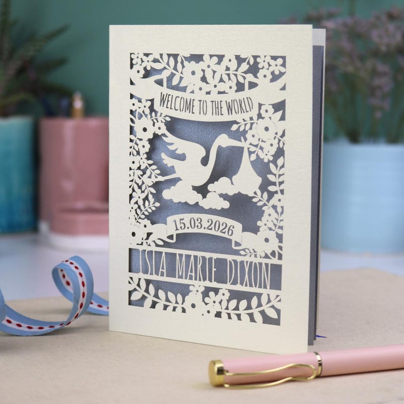 A gorgeous new baby card depicting a stork carrying a bundle surrounded with leaves, flowers and clouds. A banner saying Welcome To The World across the top,  and a second banner below with the baby's date of birth and their name under that. Shown standing on a white desk with stationery items in the fore and background. Its cut from cream cardstock and has a silver insert paper.