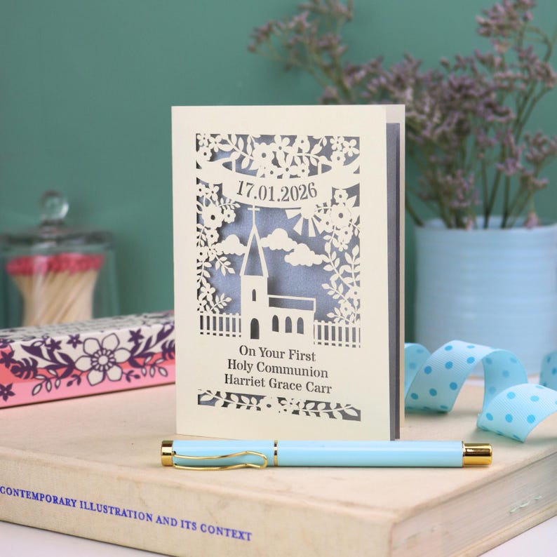 A laser cut First Holy Communion card with a church surrounded by trees, flowers, clouds and the sun. With a banner flying above the church for the  Communion date and beneath the text "On Your First Holy Communion" and the child's name. Shown standing on a white desk with stationery items, laser cut from cream cardstock with a silver insert paper