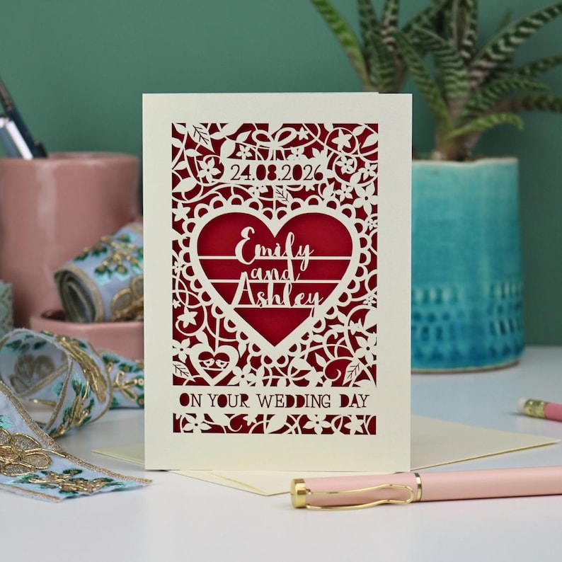 A laser cut "on your wedding day" card with floral design and cut out personalised text. Card features the wedding date, the first names of the couple in a heart shape, and the text "On your wedding day" Standing on a white desk with stationery items in the background. Cut from cream card stock with a dark red insert paper