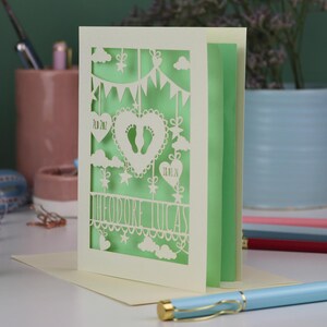 A laser cut new baby card standing on a white desk with stationery items in the background. This beautiful card is laser cut from a cream cardstock with a light green paper insert and features a heart with a pair of baby feet cut out, bunting, clouds, stars and hearts. The text under the heart is the baby's name Theodore Lucas and the left hanging heart shows the baby's weight, 7lb 2oz, and the right hanging heart shows the baby's date of birth 18/01/26