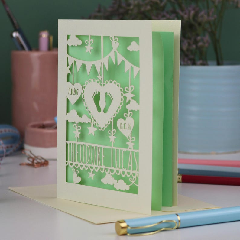 A laser cut new baby card standing on a white desk with stationery items in the background. This beautiful card is laser cut from a cream cardstock with a light green paper insert and features a heart with a pair of baby feet cut out, bunting, clouds, stars and hearts. The text under the heart is the baby's name Theodore Lucas and the left hanging heart shows the baby's weight, 7lb 2oz, and the right hanging heart shows the baby's date of birth 18/01/26