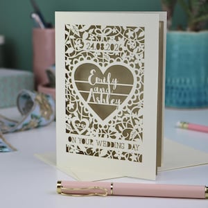A laser cut "on your wedding day" card with floral design and cut out personalised text. Card features the wedding date, the first names of the couple in a heart shape, and the text "On your wedding day" Standing on a white desk with stationery items in the background. Cut from cream card stock with a gold leaf insert paper