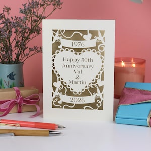 May include: A cream-colored 50th-anniversary card with gold accents. The card features a heart-shaped design with the text "Happy 50th Anniversary Val & Martin" and the years 1976 and 2026. The card is surrounded by decorative cutouts.