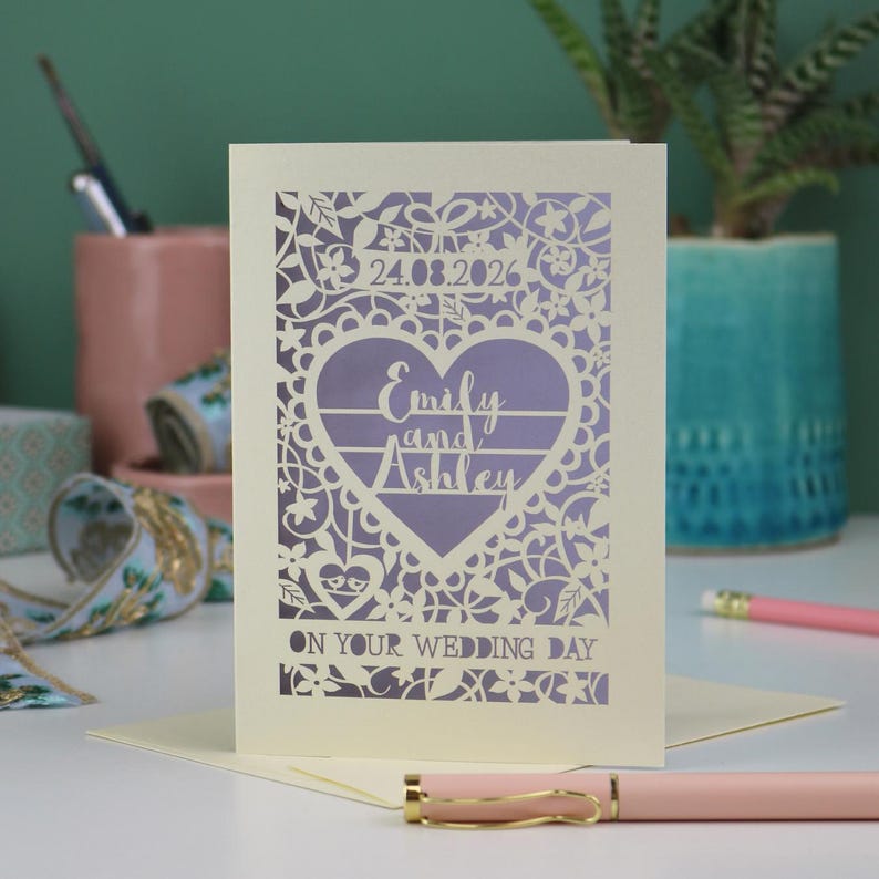 A laser cut "on your wedding day" card with floral design and cut out personalised text. Card features the wedding date, the first names of the couple in a heart shape, and the text "On your wedding day" Standing on a white desk with stationery items in the background. Cut from cream card stock with a lilac insert paper