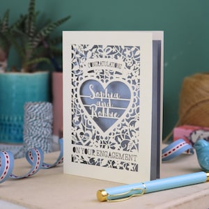 A laser cut engagement card with intricate floral cut out design, the word "Congratulations" in a banner at the top, a heart shape border with 3 lines of text, 'Name and Name', and 'On Your Engagement' in cut out lettering across the bottom of the card.
