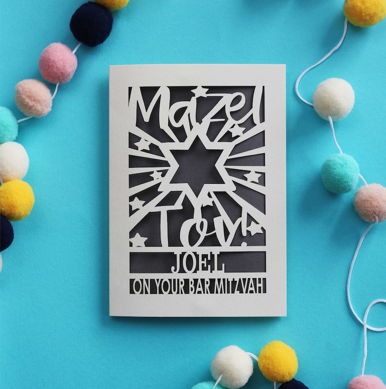 K&ouml;nnte beinhalten: Eine wei&szlig;e, lasergeschnittene Gru&szlig;karte mit einem Davidstern, dem Text "Mazel Tov!", dem Namen "JOEL" und dem Schriftzug "ON YOUR BAR MITZVAH". Die Karte hat einen dunkelgrauen Hintergrund, der durch die Ausschnitte sichtbar ist.