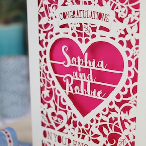 A laser cut engagement card with intricate floral cut out design, the word "Congratulations" in a banner at the top, a heart shape border with 3 lines of text, 'Name and Name', and 'On Your Engagement' in cut out lettering across the bottom of the card.