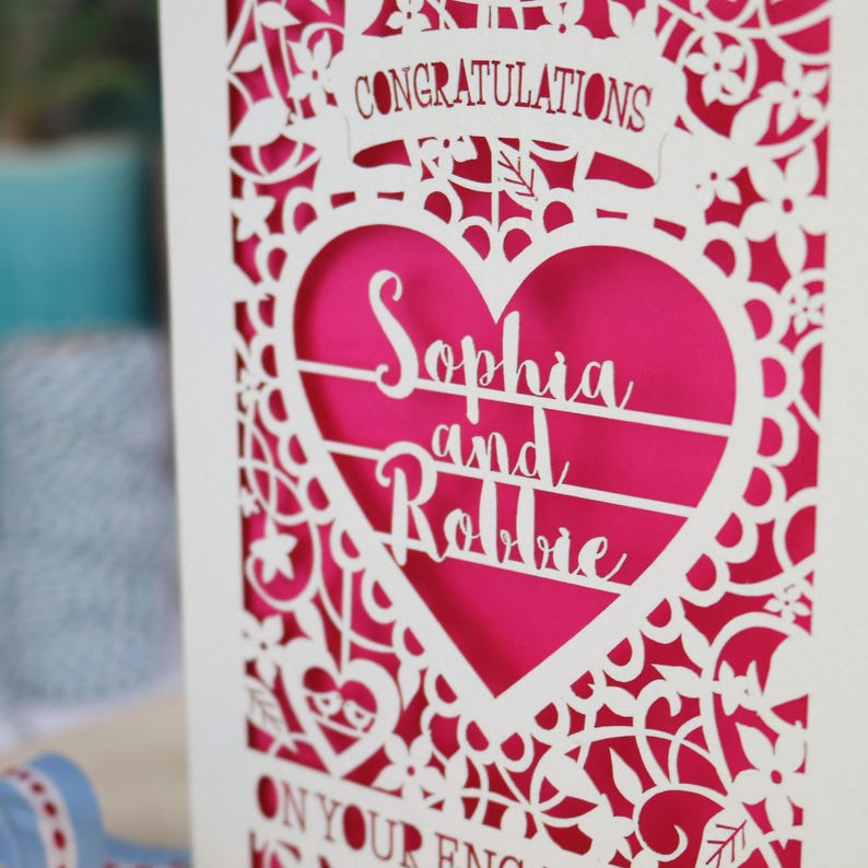 A laser cut engagement card with intricate floral cut out design, the word "Congratulations" in a banner at the top, a heart shape border with 3 lines of text, 'Name and Name', and 'On Your Engagement' in cut out lettering across the bottom of the card.