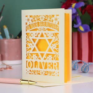 A laser cut Bar Mitzvah card with Star of David design and text "On Your Bar Mitzvah" at the top. The card is personalised with the recipient's name at the bottom and had floral and leaf design elements around the border.
