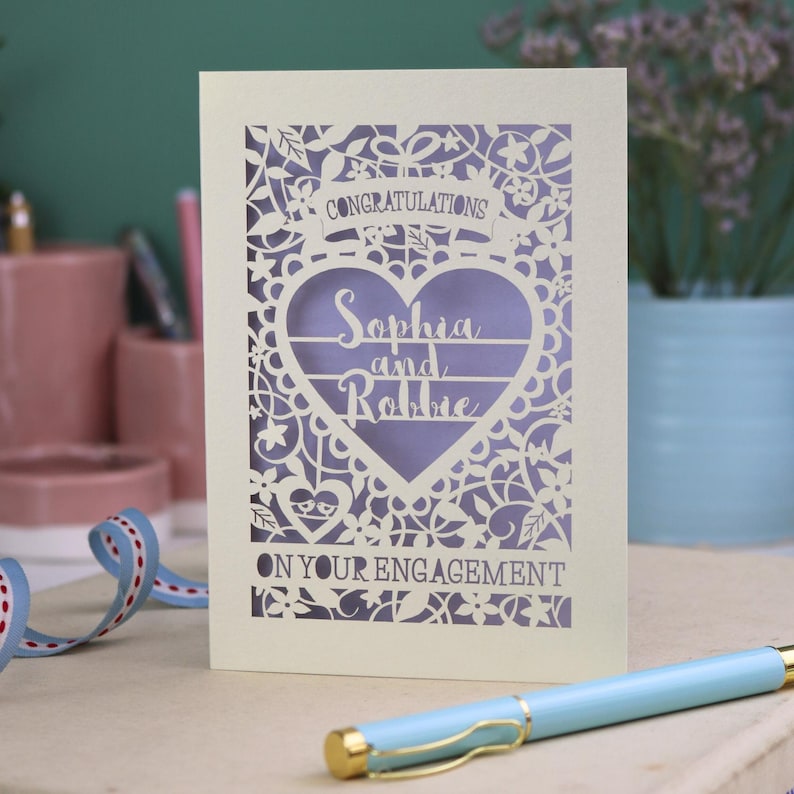 A laser cut engagement card with intricate floral cut out design, the word "Congratulations" in a banner at the top, a heart shape border with 3 lines of text, 'Name and Name', and 'On Your Engagement' in cut out lettering across the bottom of the card.