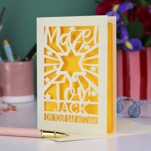 K&ouml;nnte beinhalten: Eine cremefarbene, lasergeschnittene Gru&szlig;karte mit einem Davidstern, Sonnenstrahlen und Sternen. Der Text lautet "Mazel Tov! JACK ON YOUR BAR MITZVAH". Die Karte hat einen leuchtend gelben Inneneinsatz und steht aufrecht auf einer Fl&auml;che.