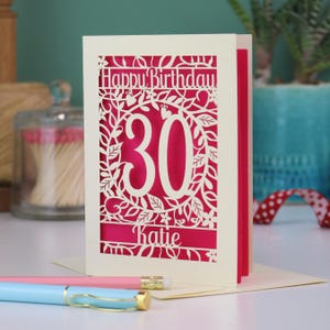 A laser cut 30th birthday card, with "Happy Birthday" text at the top, the number 30 in the centre with a floral and leaf cut out border, and personalised with a name underneath. Card is cream with a shocking pink paper insert.