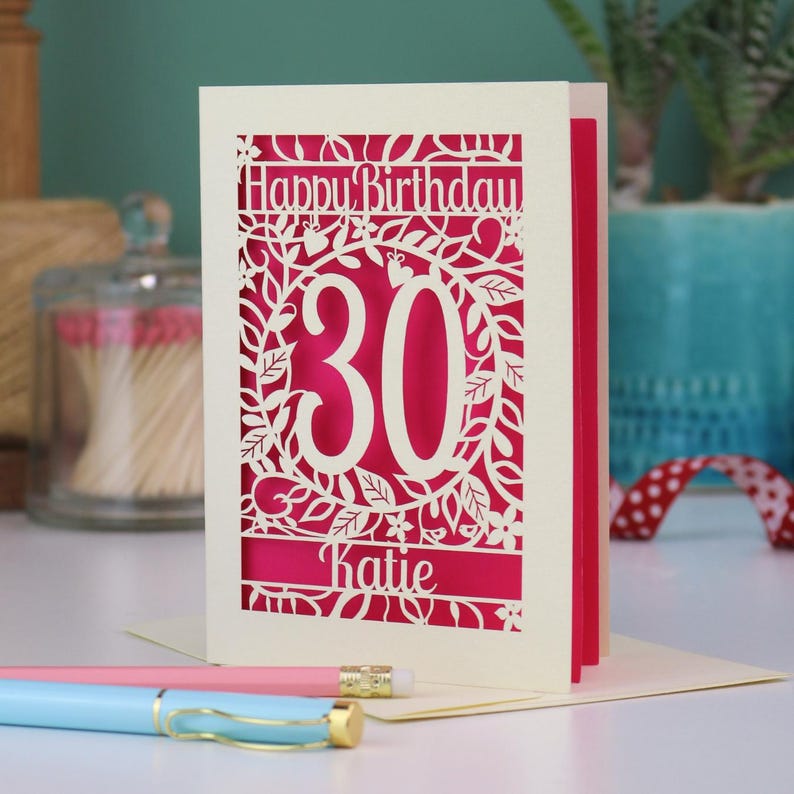 A laser cut 30th birthday card, with "Happy Birthday" text at the top, the number 30 in the centre with a floral and leaf cut out border, and personalised with a name underneath. Card is cream with a shocking pink paper insert.