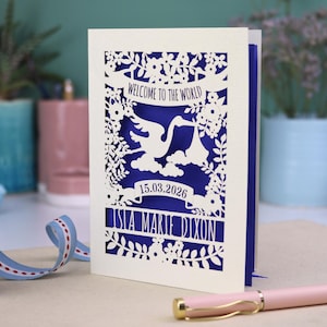 A gorgeous new baby card depicting a stork carrying a bundle surrounded with leaves, flowers and clouds. A banner saying Welcome To The World across the top,  and a second banner below with the baby's date of birth and their name under that. Shown standing on a white desk with stationery items in the fore and background. Its cut from cream cardstock and has a  violet insert paper.