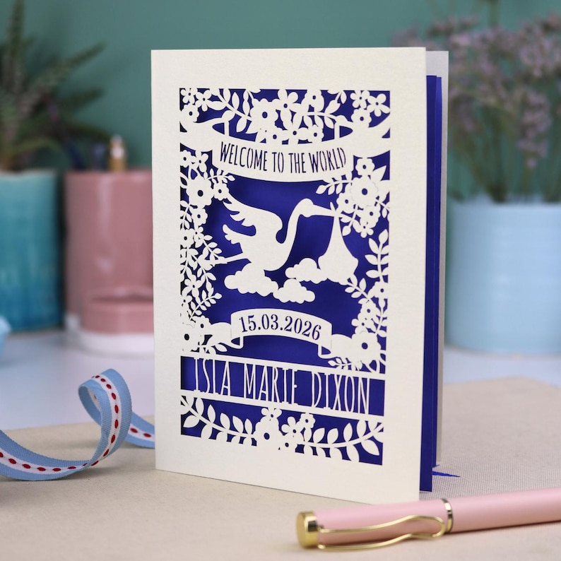 A gorgeous new baby card depicting a stork carrying a bundle surrounded with leaves, flowers and clouds. A banner saying Welcome To The World across the top,  and a second banner below with the baby's date of birth and their name under that. Shown standing on a white desk with stationery items in the fore and background. Its cut from cream cardstock and has a  violet insert paper.
