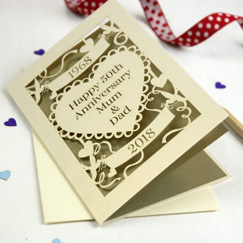 May include: A cream-colored, laser-cut anniversary card. The card reads "Happy 50th Anniversary Mum & Dad" with the years 1968 and 2018. The card has a heart-shaped design and decorative cutouts. A red and white polka dot ribbon is in the background.