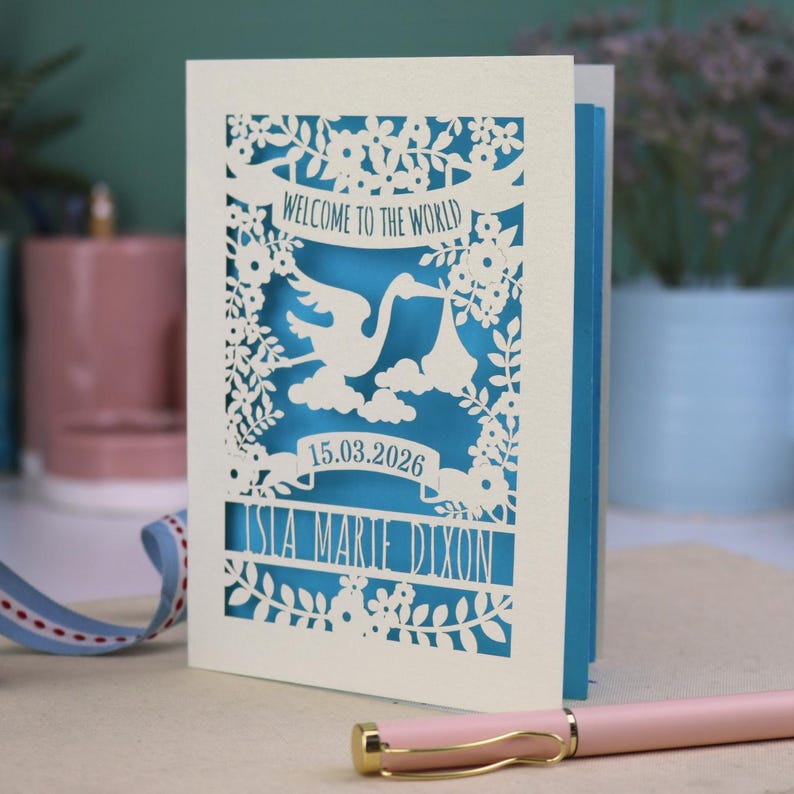A gorgeous new baby card depicting a stork carrying a bundle surrounded with leaves, flowers and clouds. A banner saying Welcome To The World across the top,  and a second banner below with the baby's date of birth and their name under that. Shown standing on a white desk with stationery items in the fore and background. Its cut from cream cardstock and has a peacock blue insert paper.