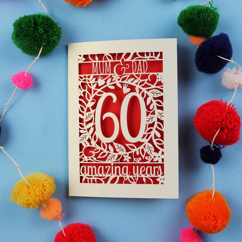 May include: A cream-colored card with a red interior, featuring a cut-out design that reads "MUM & DAD" and "60 amazing years." The card is decorated with floral patterns and the number 60. Colorful pom-poms are strung around the card on a blue background.