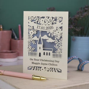 This beautiful laser cut Christening card shows a church surrounded by trees, flowers, clouds and the sun. With a banner flying above the church for the Christening date and beneath the text "On Your Christening Day" and the child's name. Shown standing on a white desk with stationery items, laser cut from cream cardstock with a silver insert paper