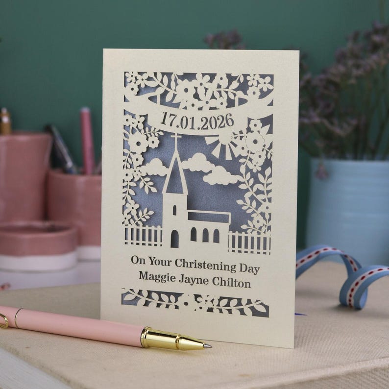 This beautiful laser cut Christening card shows a church surrounded by trees, flowers, clouds and the sun. With a banner flying above the church for the Christening date and beneath the text "On Your Christening Day" and the child's name. Shown standing on a white desk with stationery items, laser cut from cream cardstock with a silver insert paper