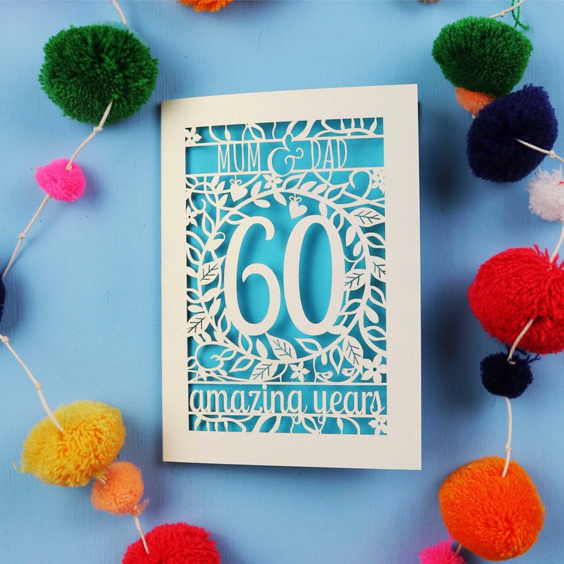 May include: A white card with intricate cut-out designs, featuring the words "MUM & DAD" and "60 amazing years" on a turquoise background. The card is decorated with floral patterns and surrounded by colorful pom-pom garland.