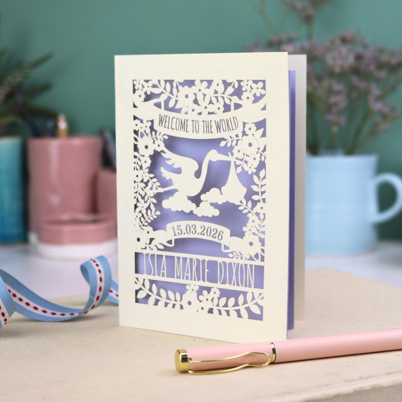 A personalised laser cut new baby card depicting a stork carrying a baby bundle surrounded with leaves, flowers and clouds. A banner saying Welcome To The World across the top,  and a second banner below with the baby's date of birth and their name under that. Shown standing on a white desk with stationery items in the fore and background. Its cut from cream cardstock and has a lilac insert paper.