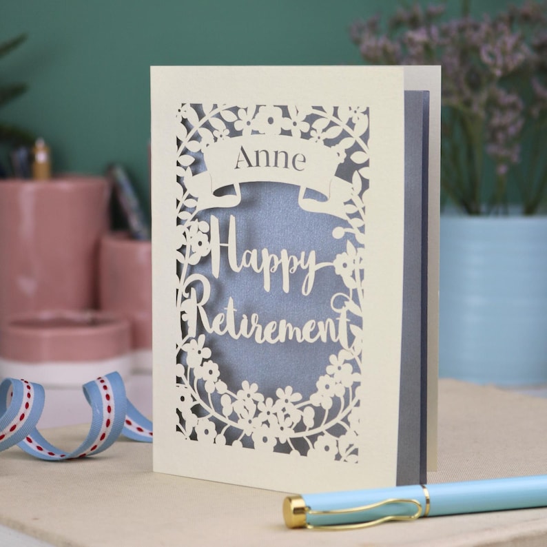 This lovely laser cut retirement card is shown standing on a white desk with a pen and ceramics in the background. It has the words Happy Retirement cut in a script font, has a banner above for the retiree's name and a floral decorative border. It is cut from cream cardstock and has a silver insert paper
