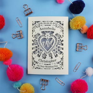 A personalised laser cut wedding card with floral filigree papercut design. Card has a space for the date at the top, a banner with a name, a heart shape with the word 'and' and another banner for the second person's name. Card says on your wedding day at the bottom.