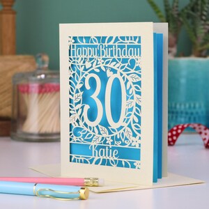 A laser cut 30th birthday card, with "Happy Birthday" text at the top, the number 30 in the centre with a floral and leaf cut out border, and personalised with a name underneath. Card is cream with a peacock blue paper insert.