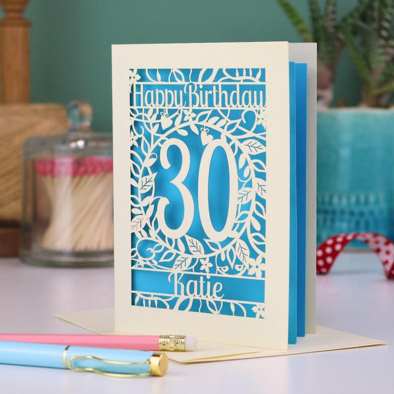 A laser cut 30th birthday card, with "Happy Birthday" text at the top, the number 30 in the centre with a floral and leaf cut out border, and personalised with a name underneath. Card is cream with a peacock blue paper insert.