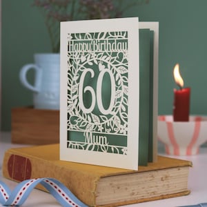 A laser cut flower birthday card with cut out floral design and "Happy Birthday" text. The birthday card is personalised with a cut out name and the age in the centre, surrounded by leaves and flowers
