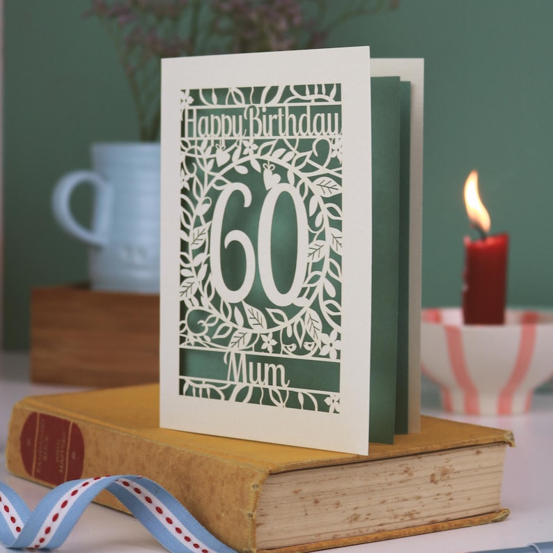 A laser cut flower birthday card with cut out floral design and "Happy Birthday" text. The birthday card is personalised with a cut out name and the age in the centre, surrounded by leaves and flowers