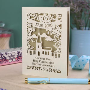 This beautiful laser cut First Holy Communion card shows a church surrounded by trees, flowers, clouds and the sun. With a banner flying above the church for the  Communion date and beneath the text "On Your First Holy Communion" and the child's name. Shown standing on a white desk with stationery items, laser cut from cream cardstock with a gold leaf insert paper