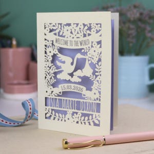A gorgeous new baby card depicting a stork carrying a bundle surrounded with leaves, flowers and clouds. A banner saying Welcome To The World across the top,  and a second banner below with the baby's date of birth and their name under that. Shown standing on a white desk with stationery items in the fore and background. Its cut from cream cardstock and has a lilac insert paper.