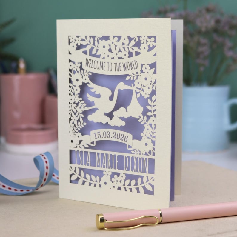 A gorgeous new baby card depicting a stork carrying a bundle surrounded with leaves, flowers and clouds. A banner saying Welcome To The World across the top,  and a second banner below with the baby's date of birth and their name under that. Shown standing on a white desk with stationery items in the fore and background. Its cut from cream cardstock and has a lilac insert paper.