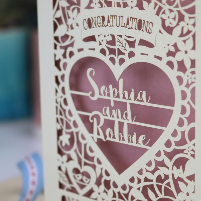 A laser cut engagement card with intricate floral cut out design, the word "Congratulations" in a banner at the top, a heart shape border with 3 lines of text, 'Name and Name', and 'On Your Engagement' in cut out lettering across the bottom of the card.