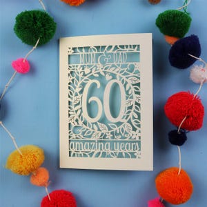 May include: A white, laser-cut card with the words "MUM & DAD", the number "60", and "amazing years". The card is decorated with floral and leaf patterns. Colorful pom-pom garland surrounds the card on a blue background.