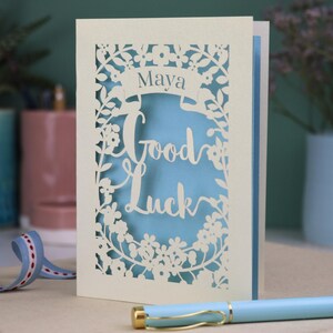 This floral Good Luck card is shown standing on a book with a pen, ribbon and ceramics in the background. It shows the text Good Luck in the centre of the card with a banner above showing the name Maya. It is laser cut from cream card with a light blue insert paper
