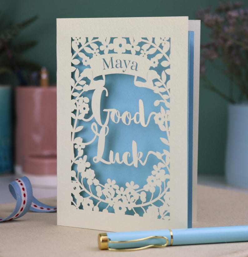 This floral Good Luck card is shown standing on a book with a pen, ribbon and ceramics in the background. It shows the text Good Luck in the centre of the card with a banner above showing the name Maya. It is laser cut from cream card with a light blue insert paper