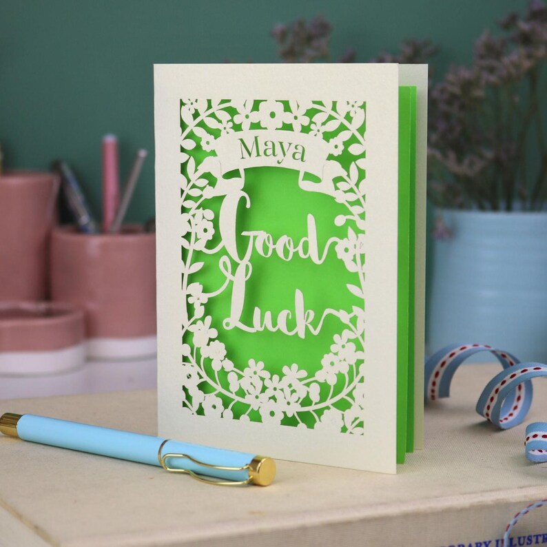 This floral Good Luck card is shown standing on a book with a pen, ribbon and ceramics in the background. It shows the text Good Luck in the centre of the card with a banner above showing the name Maya. It is laser cut from cream card with a bright green insert paper