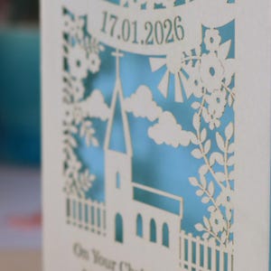 A close up detail of this beautiful laser cut Christening card shows a church surrounded by trees, flowers, clouds and the sun. With a banner flying above the church for the Christening date and beneath the text "On Your Christening Day" and the child's name. Shown standing on a white desk with stationery items, laser cut from cream cardstock with a light blue insert paper