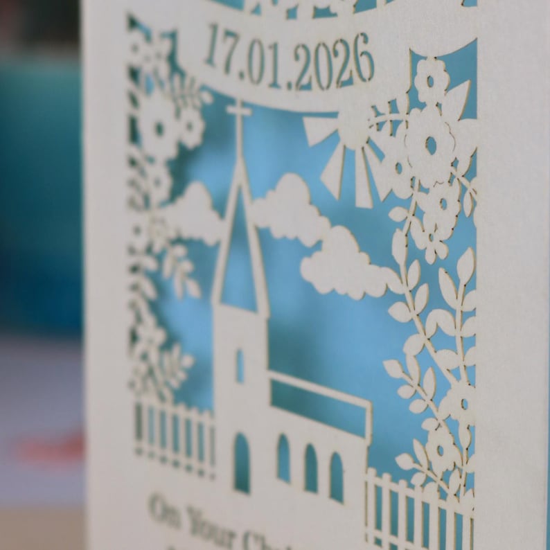 A close up detail of this beautiful laser cut Christening card shows a church surrounded by trees, flowers, clouds and the sun. With a banner flying above the church for the Christening date and beneath the text "On Your Christening Day" and the child's name. Shown standing on a white desk with stationery items, laser cut from cream cardstock with a light blue insert paper