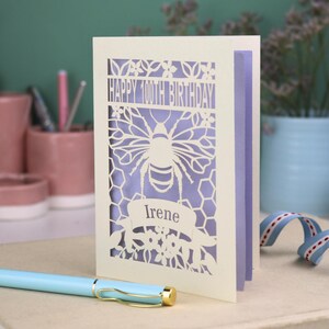 Peut inclure: Une carte de v&oelig;ux d&eacute;coup&eacute;e au laser de couleur cr&egrave;me avec une abeille, des motifs en nid d'abeille et des fleurs. Le texte indique "HAPPY 100TH BIRTHDAY" en haut et "Irene" sur une banni&egrave;re sous l'abeille. La carte a un insert violet clair.