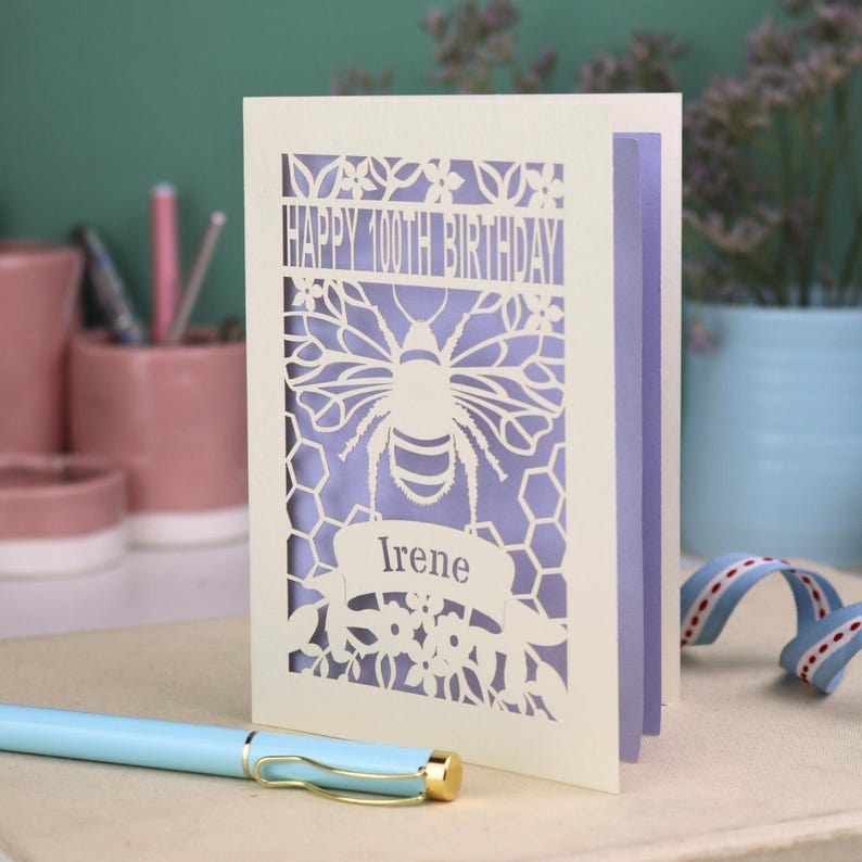 Peut inclure: Une carte de v&oelig;ux d&eacute;coup&eacute;e au laser de couleur cr&egrave;me avec une abeille, des motifs en nid d'abeille et des fleurs. Le texte indique "HAPPY 100TH BIRTHDAY" en haut et "Irene" sur une banni&egrave;re sous l'abeille. La carte a un insert violet clair.