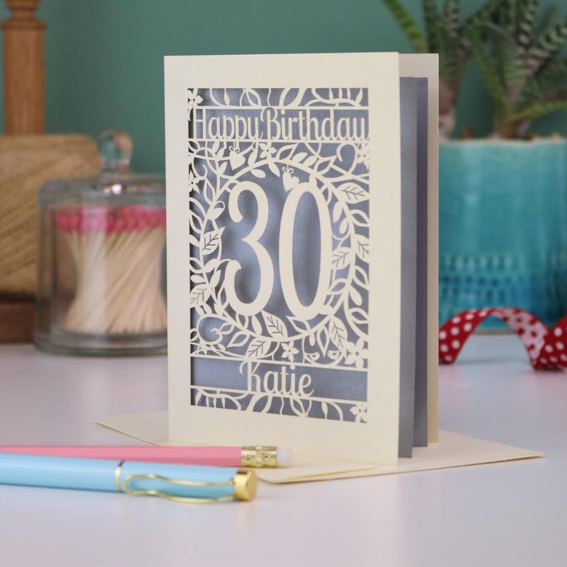 A laser cut 30th birthday card, with "Happy Birthday" text at the top, the number 30 in the centre with a floral and leaf cut out border, and personalised with a name underneath. Card is cream with a silver paper insert.