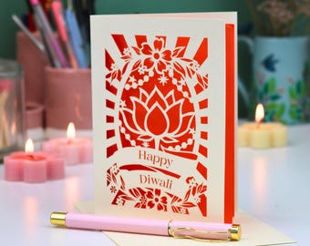 Op de afbeelding: Een crèmekleurige wenskaart met een felrood lasergesneden ontwerp van een lotusbloem omringd door bloemmotieven en zonnestralen. De tekst Happy Diwali staat in het rood op de voorzijde. Een lichtroze pen ligt horizontaal voor de kaart.