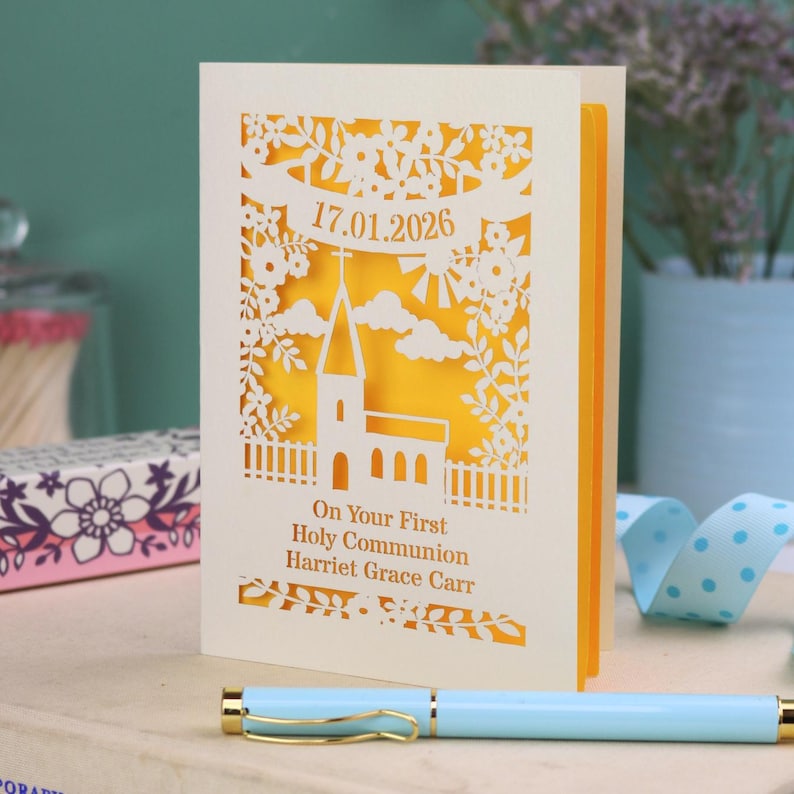This beautiful laser cut First Holy Communion card shows a church surrounded by trees, flowers, clouds and the sun. With a banner flying above the church for the  Communion date and beneath the text "On Your First Holy Communion" and the child's name. Shown standing on a white desk with stationery items, laser cut from cream cardstock with a sunshine yellow insert paper